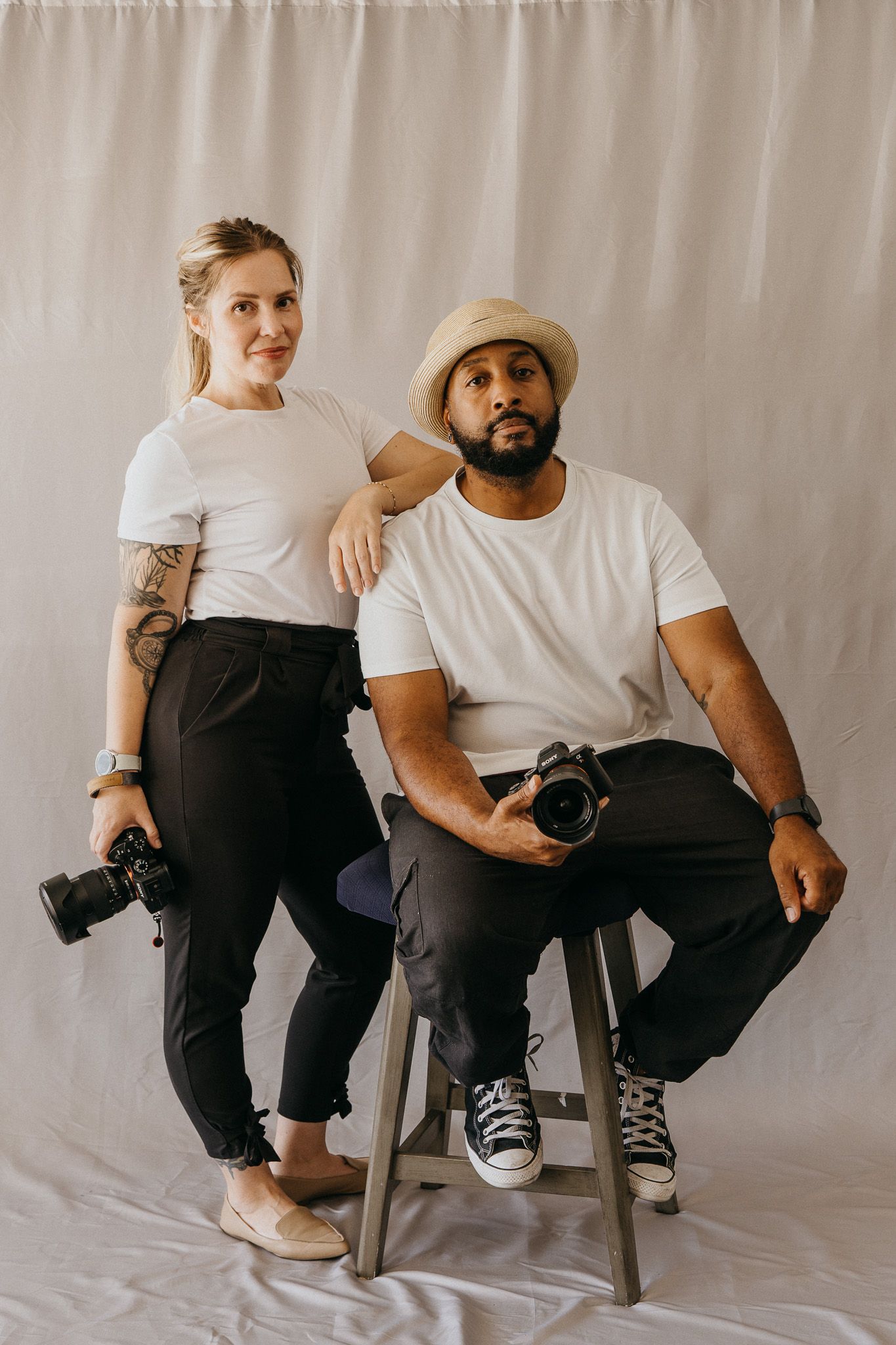 Two photographers in white t-shirts pose in a studio, holding cameras, one seated on a stool.