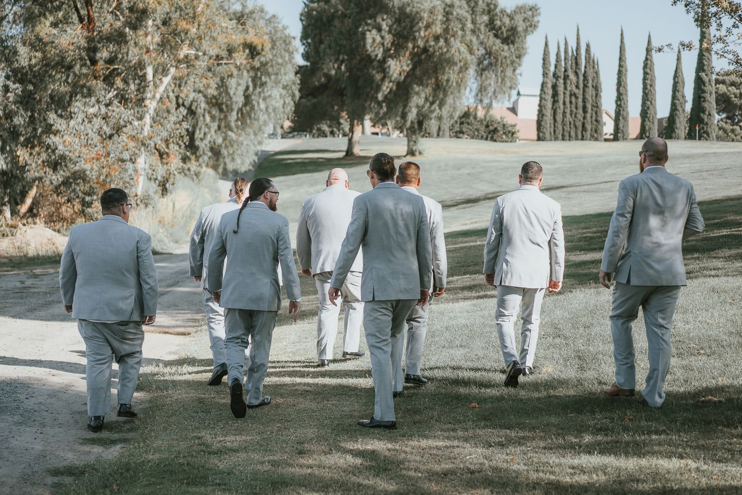 Groomsmen in matching light gray suits walking together on a path at an outdoor wedding venue.