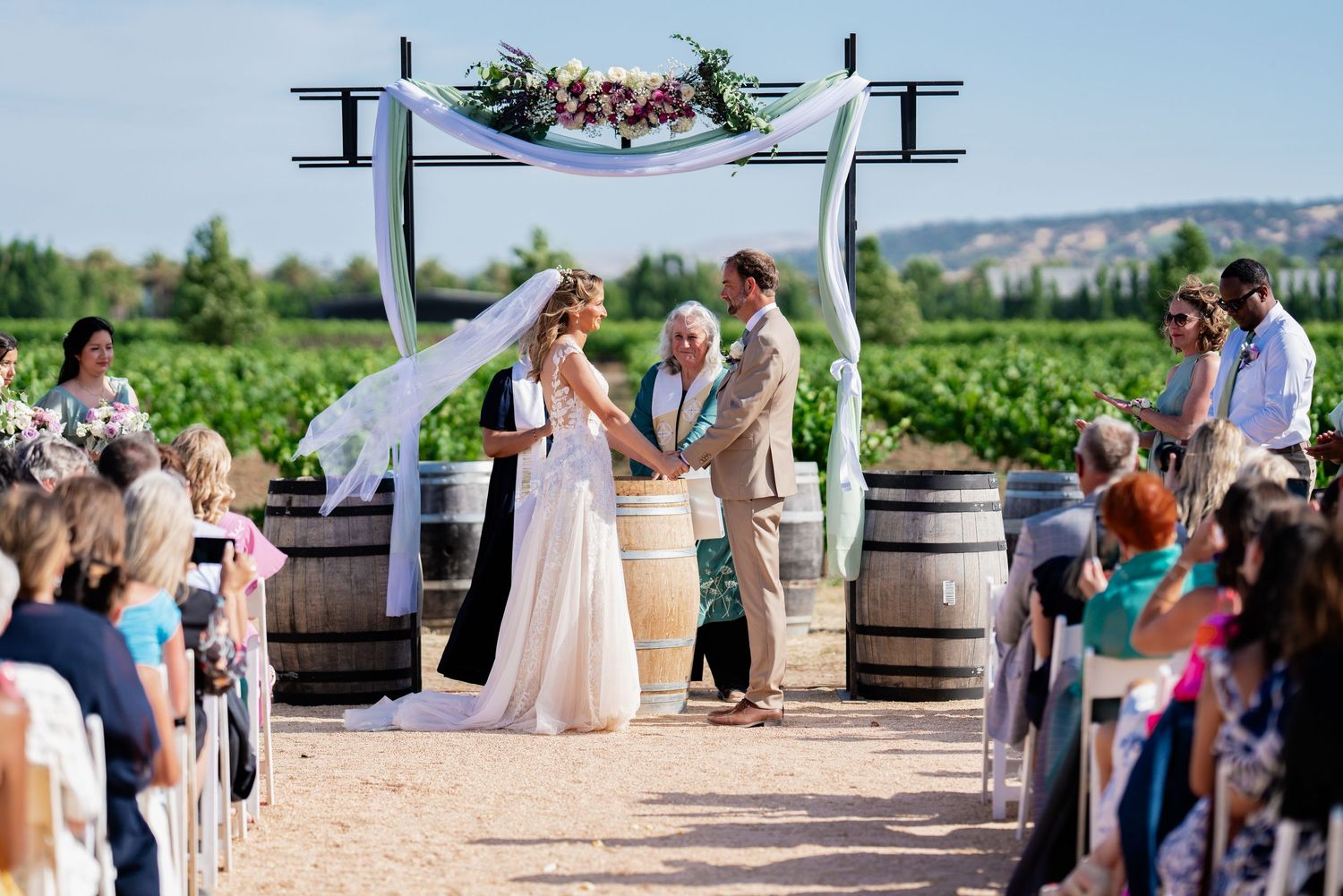 Outdoor vineyard wedding ceremony with couple exchanging vows under floral arch flanked by wine barrels.