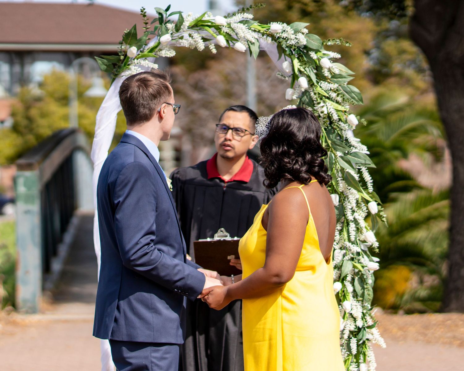 Couple exchanging vows under floral arch with officiant at outdoor wedding ceremony.