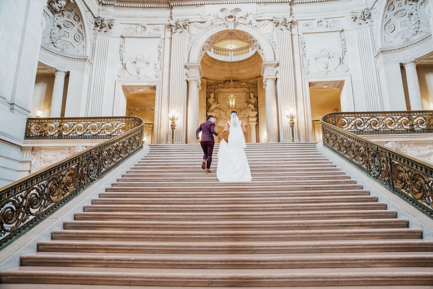 Bride and groom ascending grand marble staircase at San Francisco City Hall during elegant wedding ceremony.