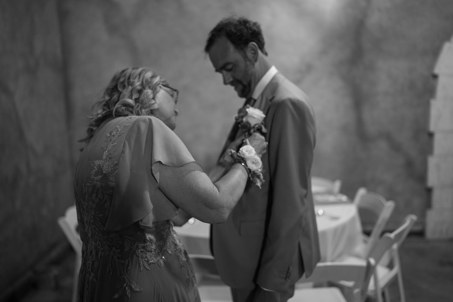 Black and white photo of a woman pinning a boutonniere on a mans suit jacket at a wedding ceremony.