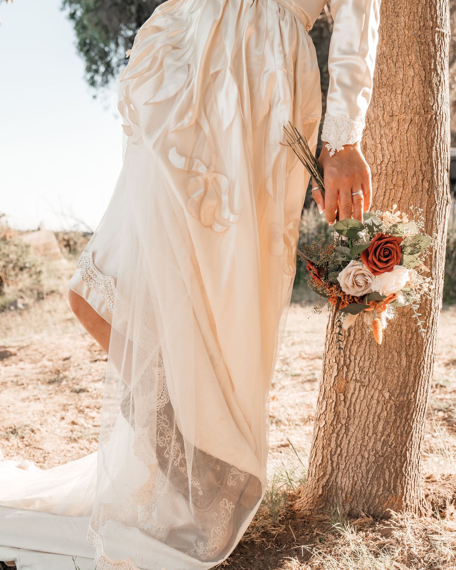 Bride in elegant white lace wedding gown holding a rustic bouquet of red and cream roses beside a tree outdoors.