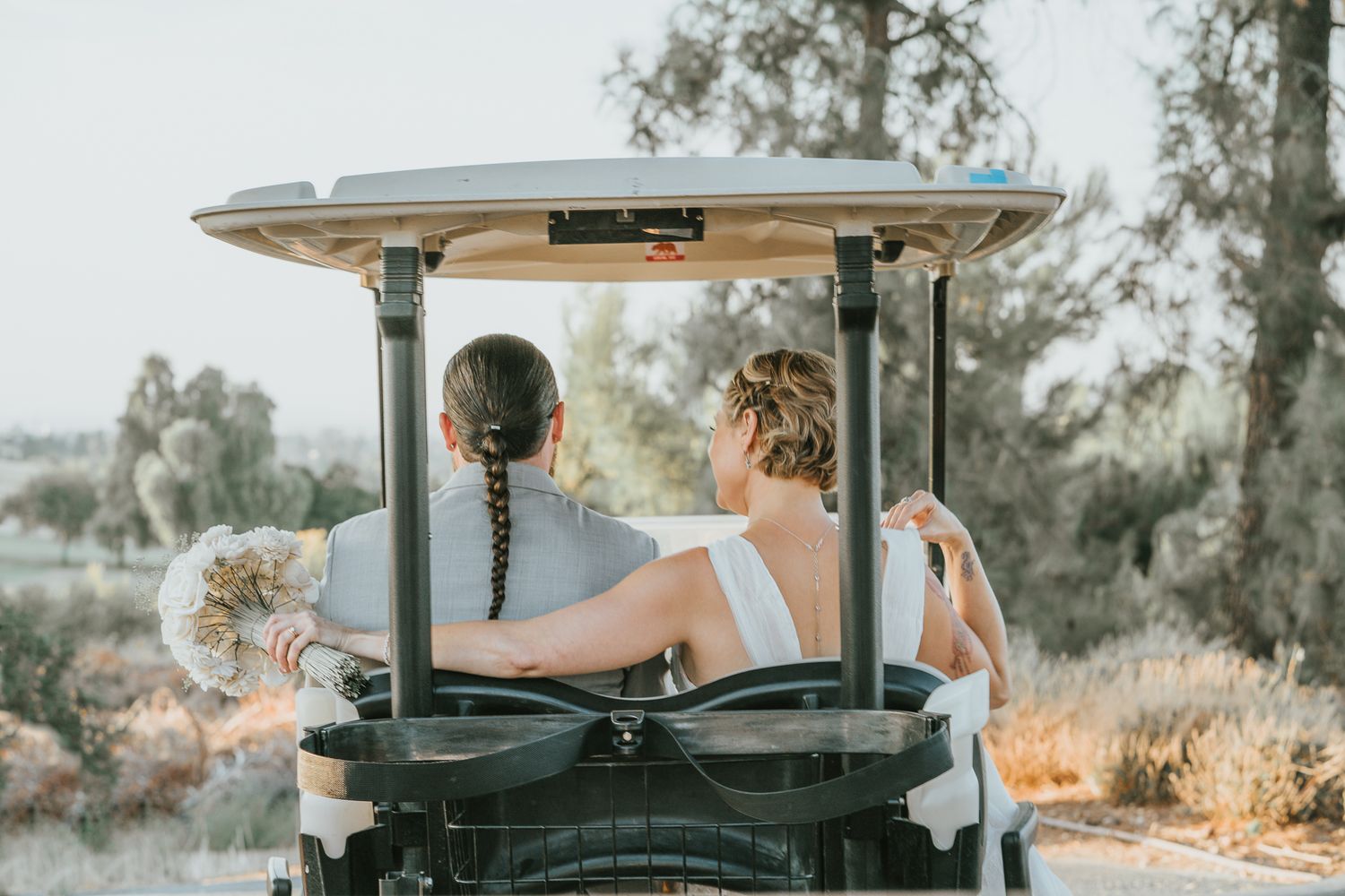 Two brides in white dresses ride golf cart through scenic outdoor wedding venue from behind.