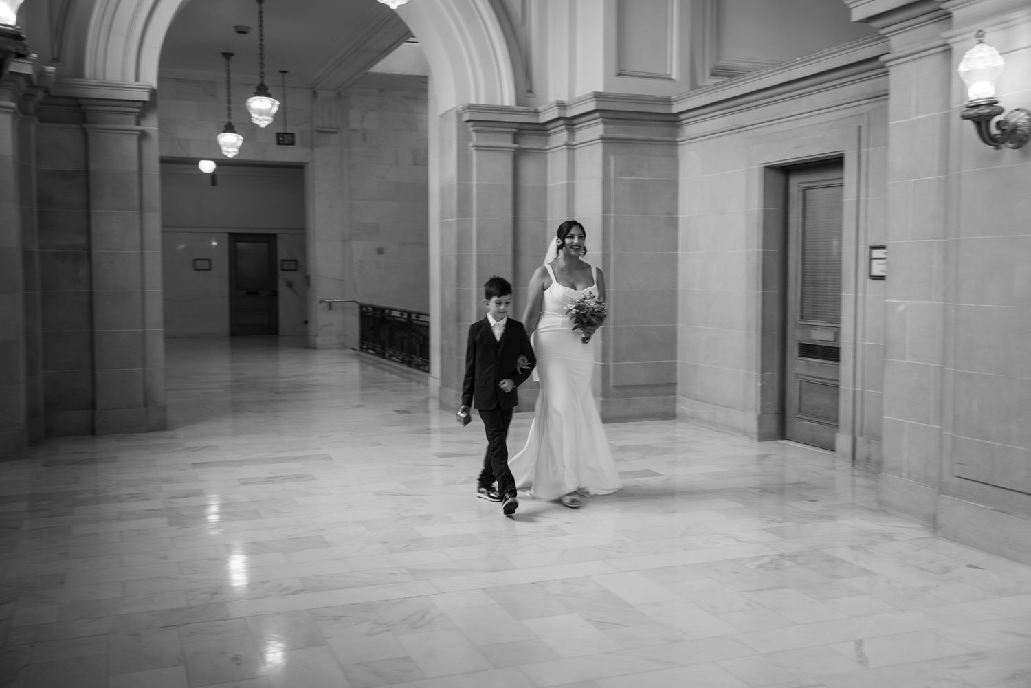 Bride in white gown walks with young boy in suit through elegant marble hallway in black and white wedding photo.