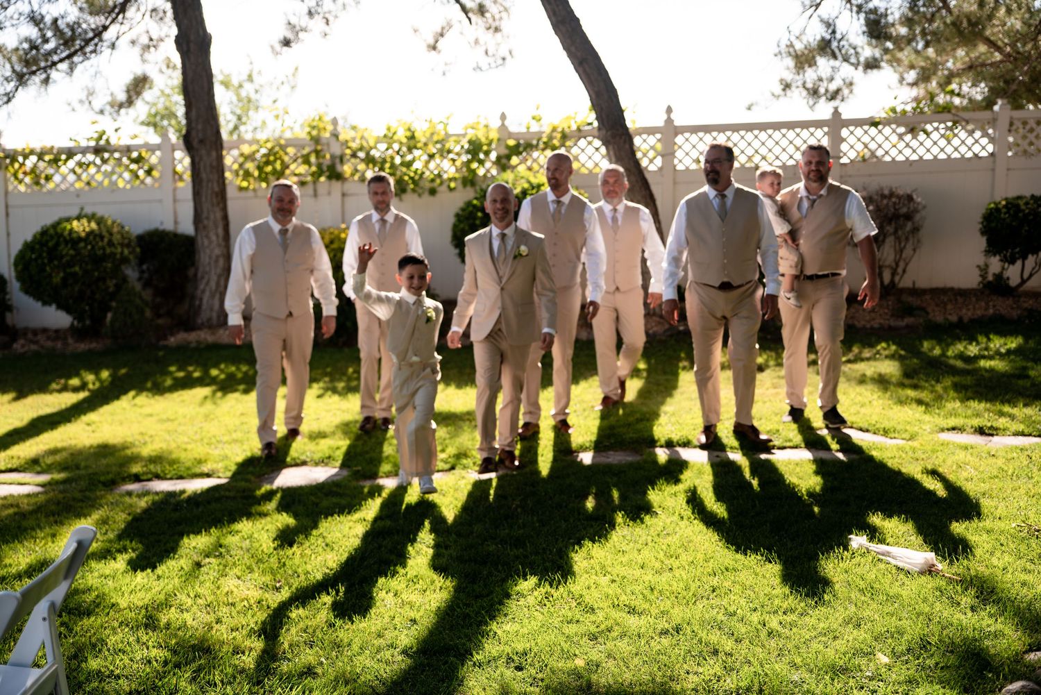 Groomsmen in tan suits walk across green lawn at outdoor wedding venue on a sunny day.