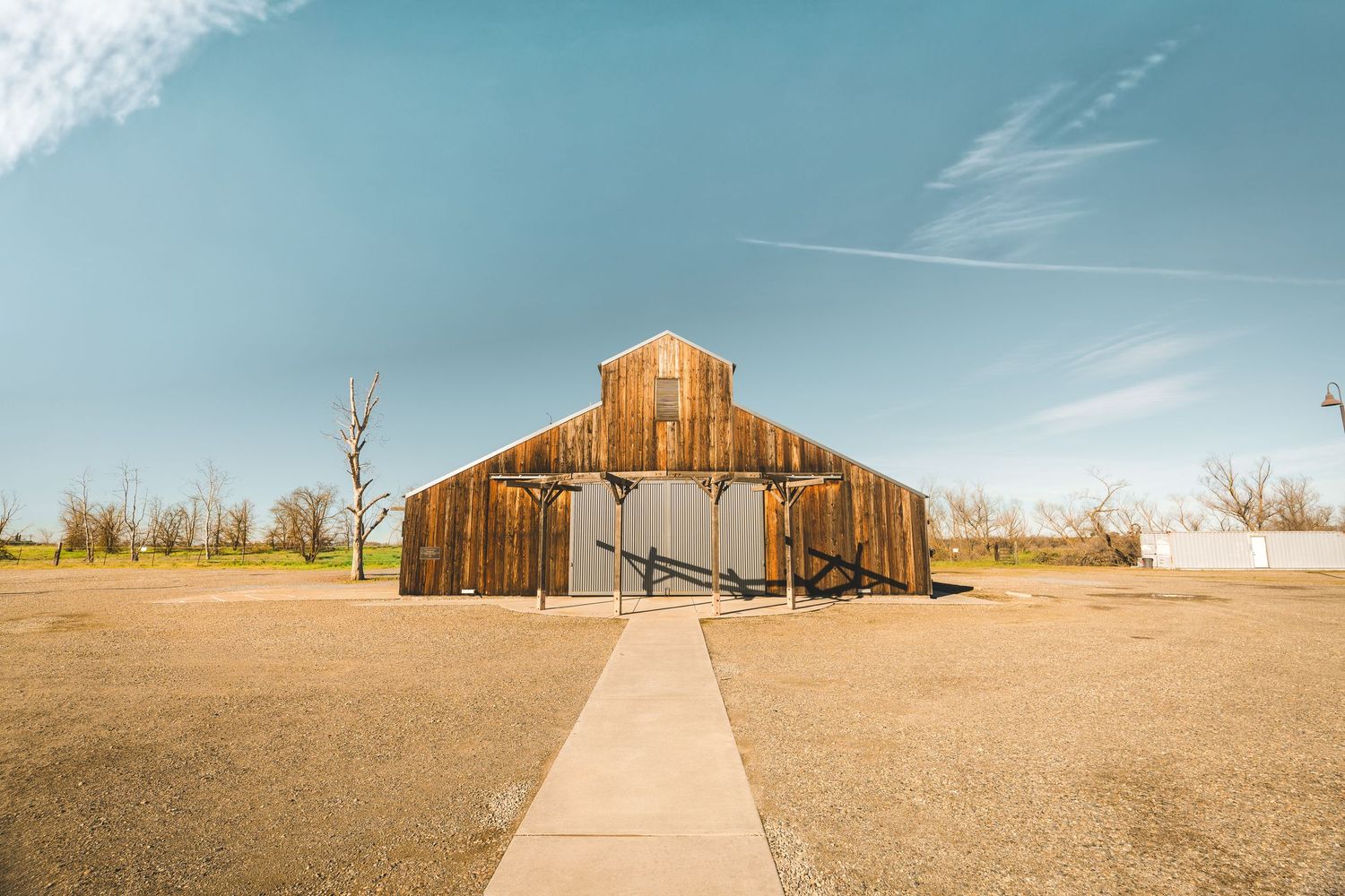 Rustic wooden barn with a concrete pathway leading to its entrance under a clear blue sky in a rural setting.