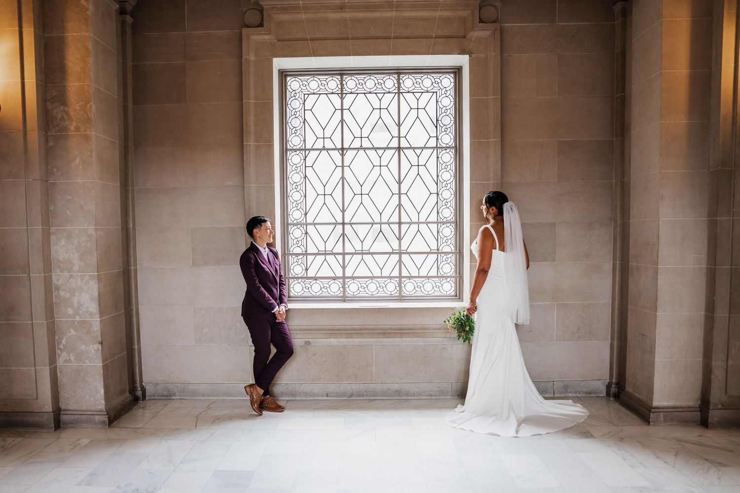 Bride and groom share a tender first look moment in an elegant stone hall with ornate lattice windows.