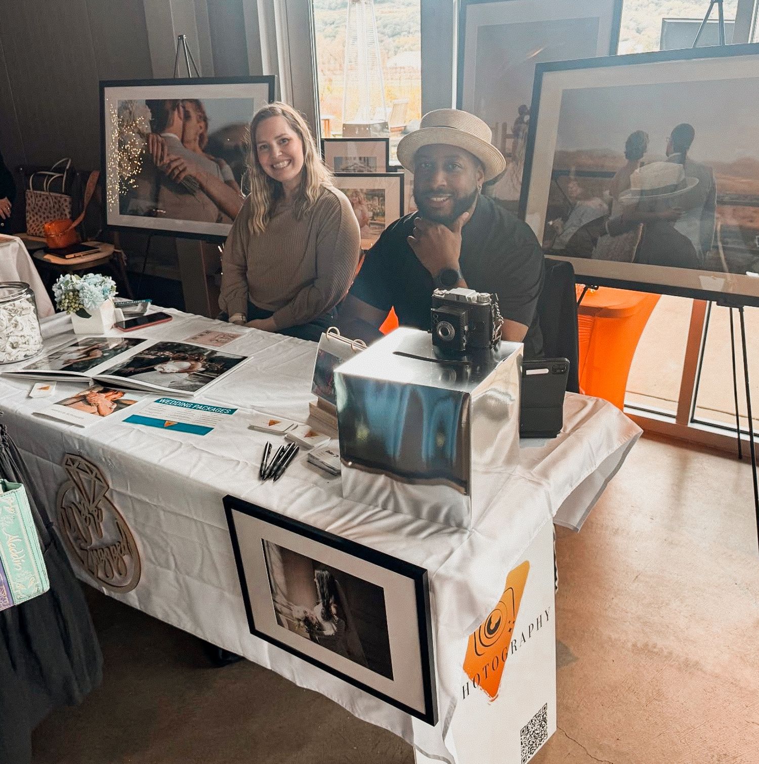 Two people smile at an art fair booth displaying framed photography prints and a vintage camera on a white tablecloth.