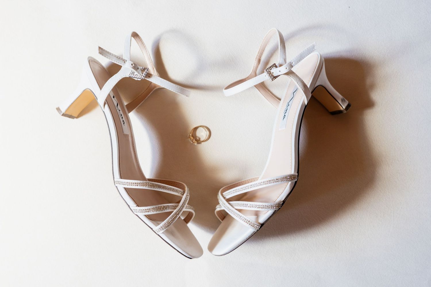 White strappy bridal heels with a gold wedding ring placed between them on a white background.
