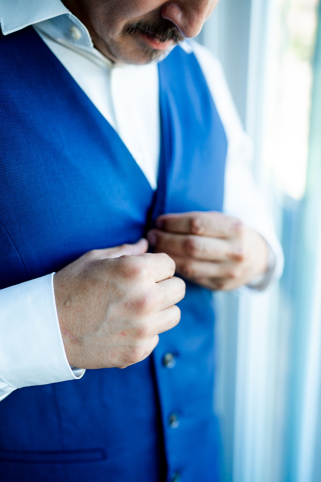 Groom in blue vest and white dress shirt buttoning his waistcoat while getting ready for wedding.