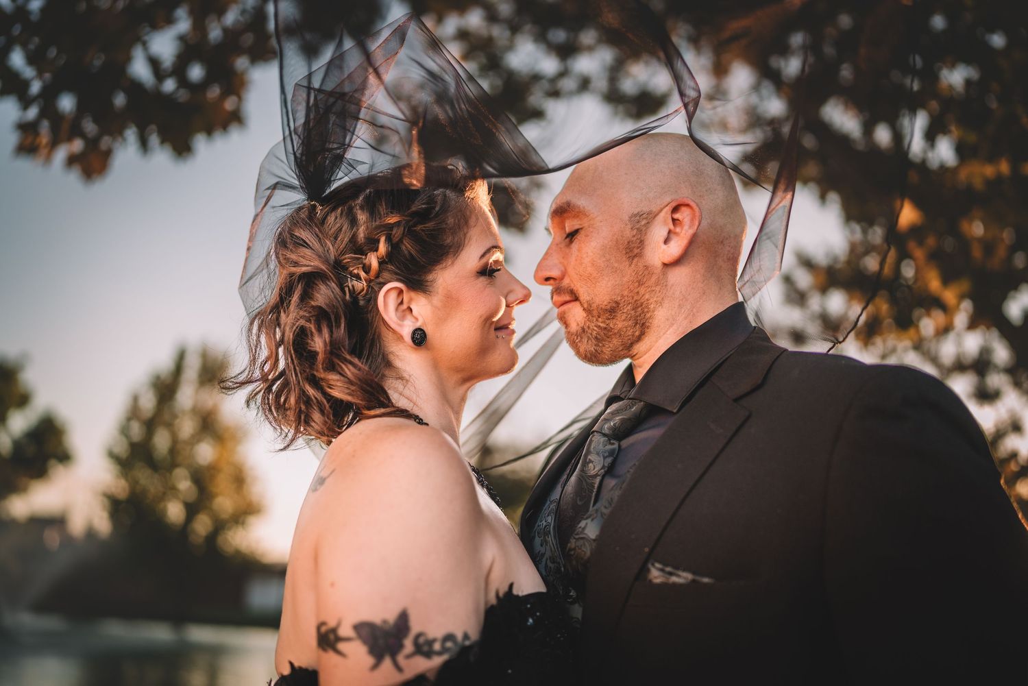 Romantic couple in wedding attire sharing an intimate moment outdoors at sunset, bride with fascinator and groom in suit.