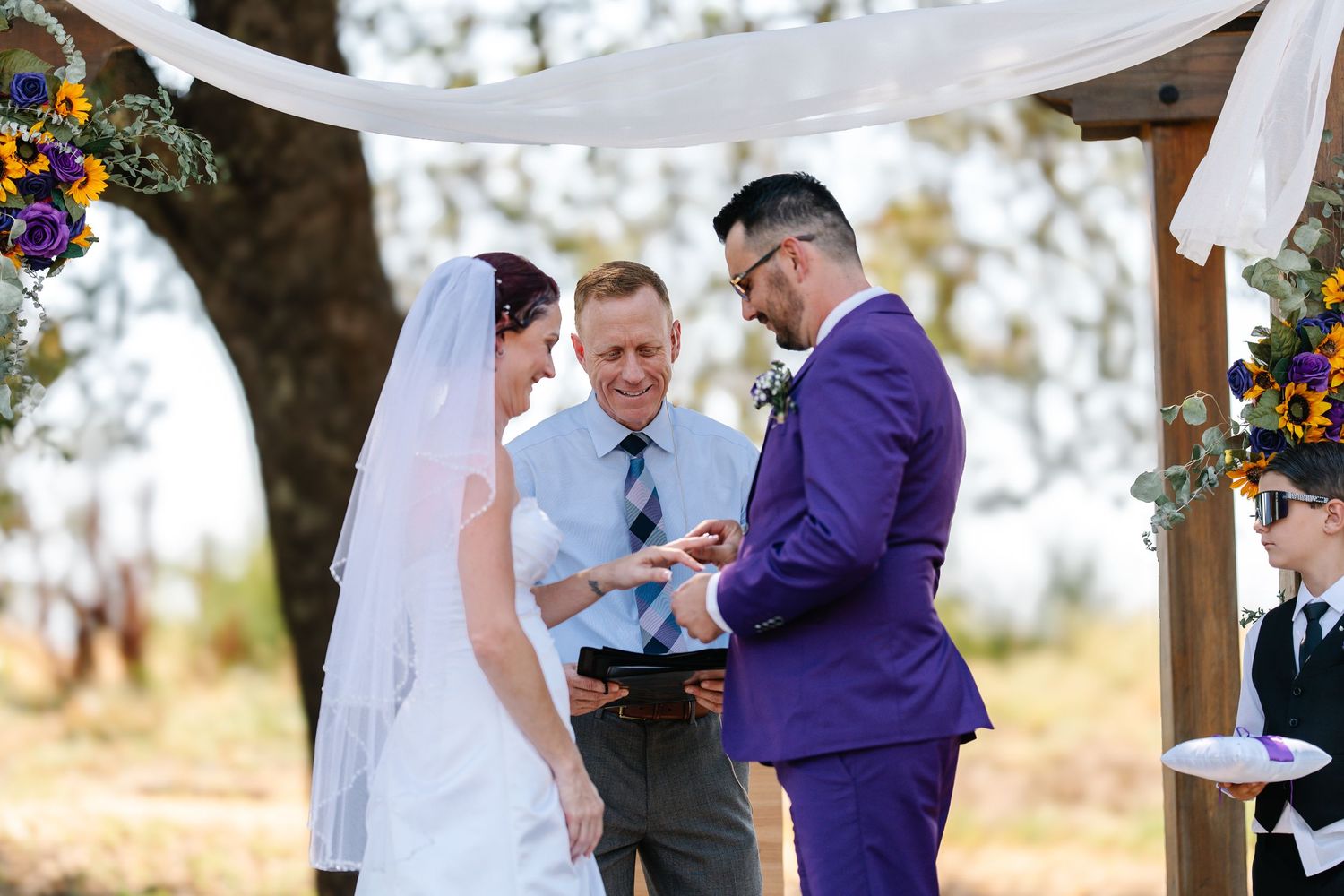 Groom in purple suit exchanges rings with bride during outdoor wedding ceremony under wooden arch.