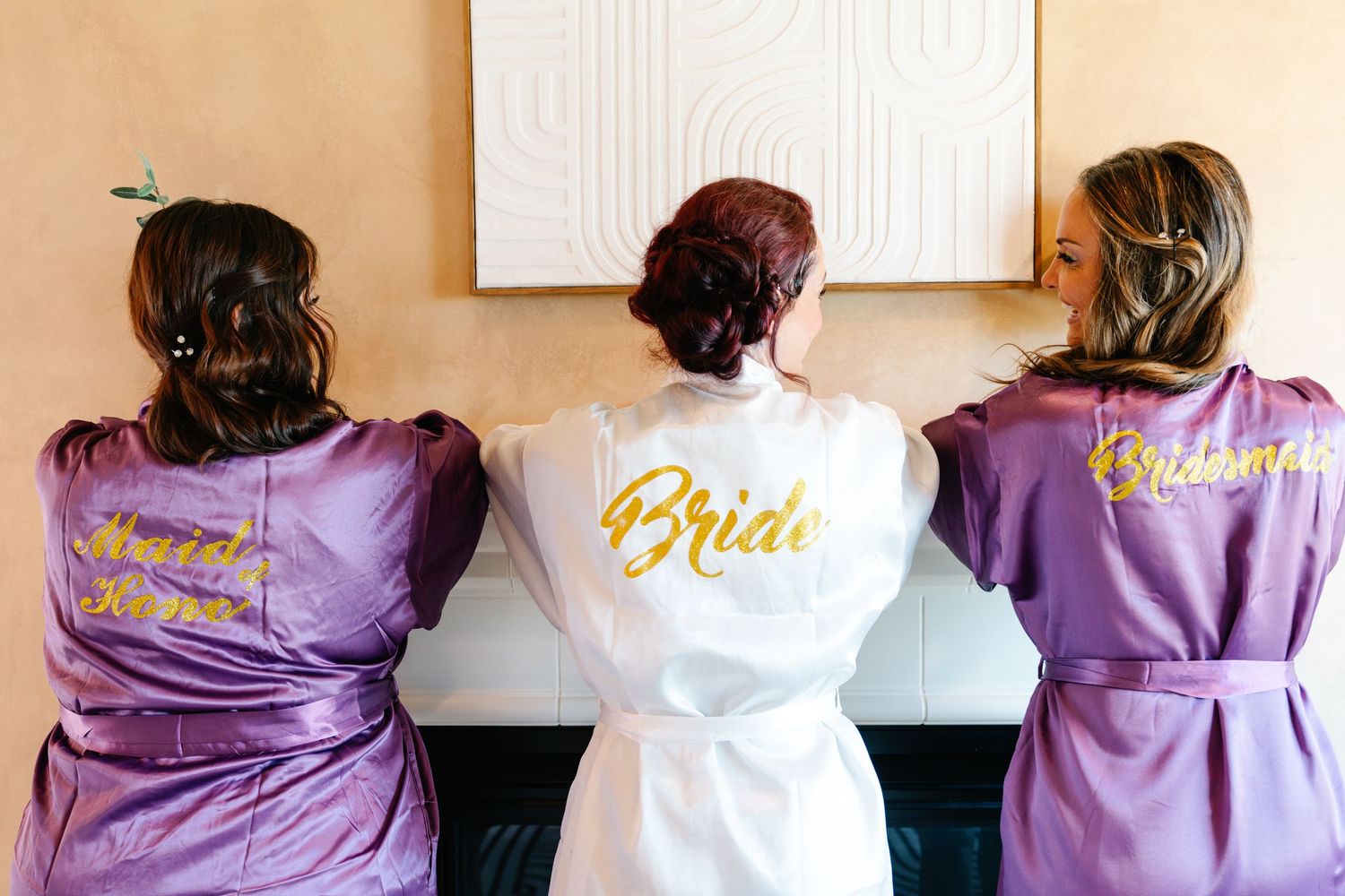 Bride and two bridesmaids wearing personalized satin robes with gold script, viewed from behind.