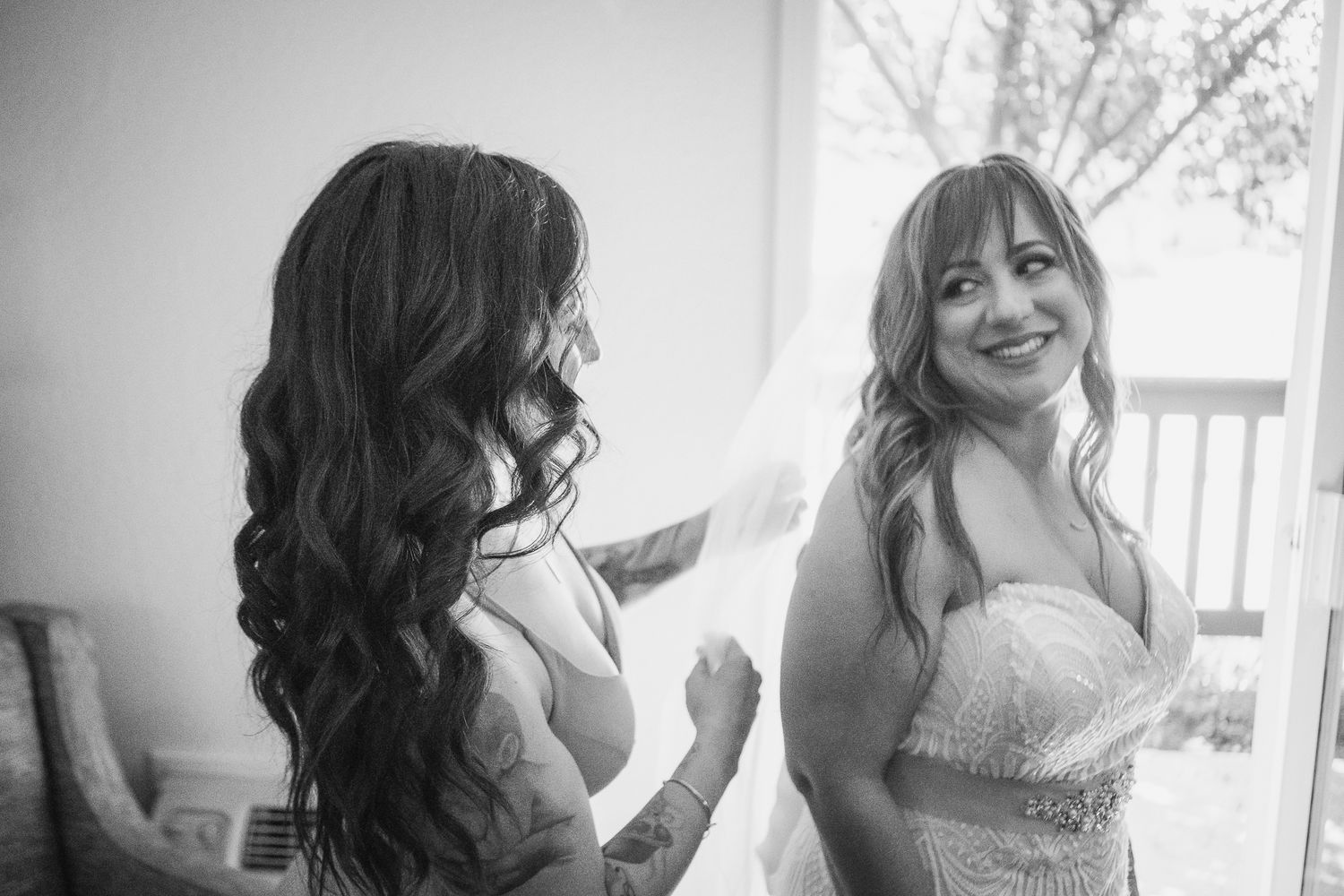 Black and white photo of a bride in a strapless gown with veil, smiling with her bridesmaid during wedding preparations.