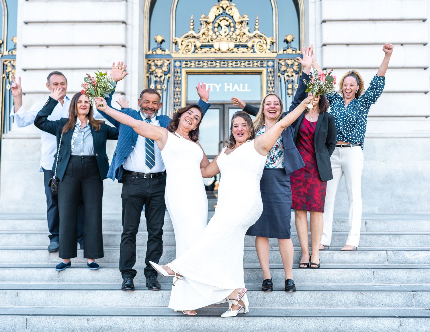 Two brides in white dresses celebrate with wedding party on San Francisco City Hall steps.