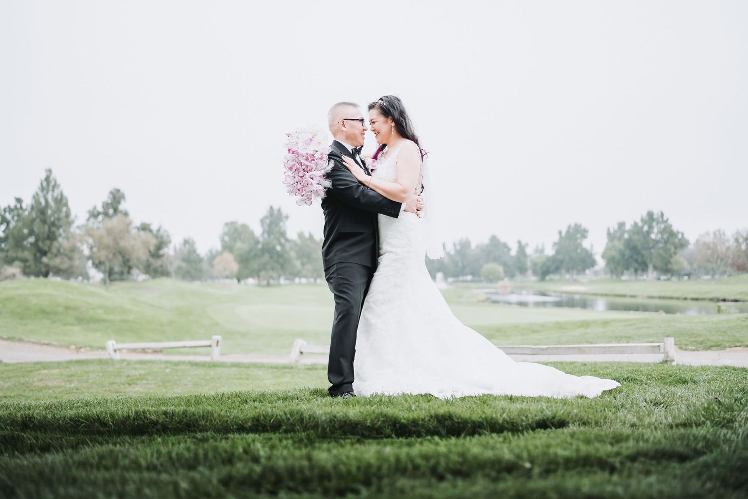 Bride and groom embracing on lush green golf course, bride holding pink bouquet in white gown, groom in black tuxedo.