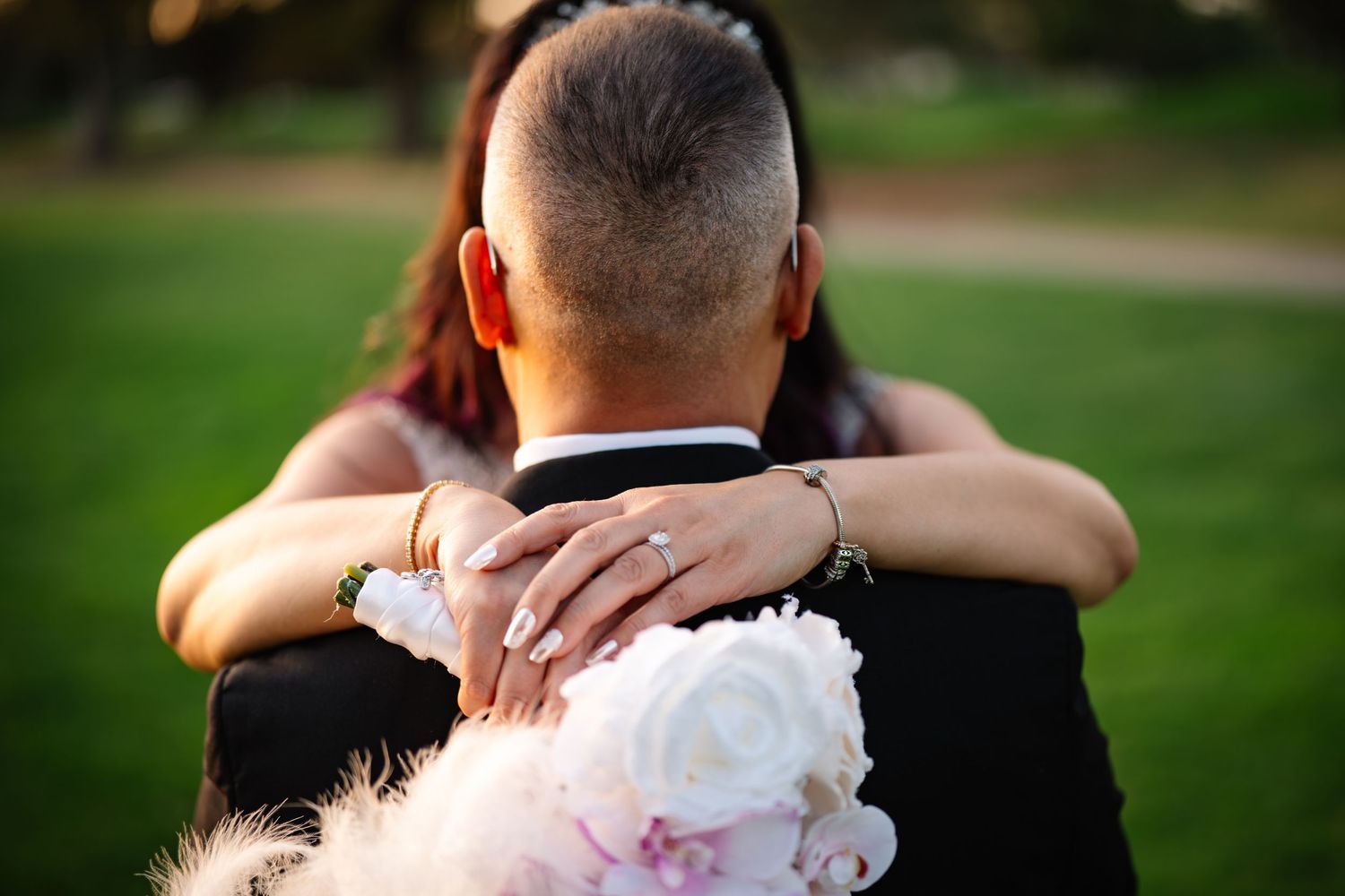 Bride embracing groom from behind, showing wedding ring and holding white floral bouquet outdoors.