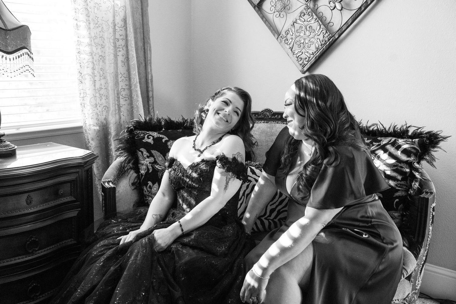 Two women in elegant black gowns laugh together on a vintage sofa in a black and white photo.