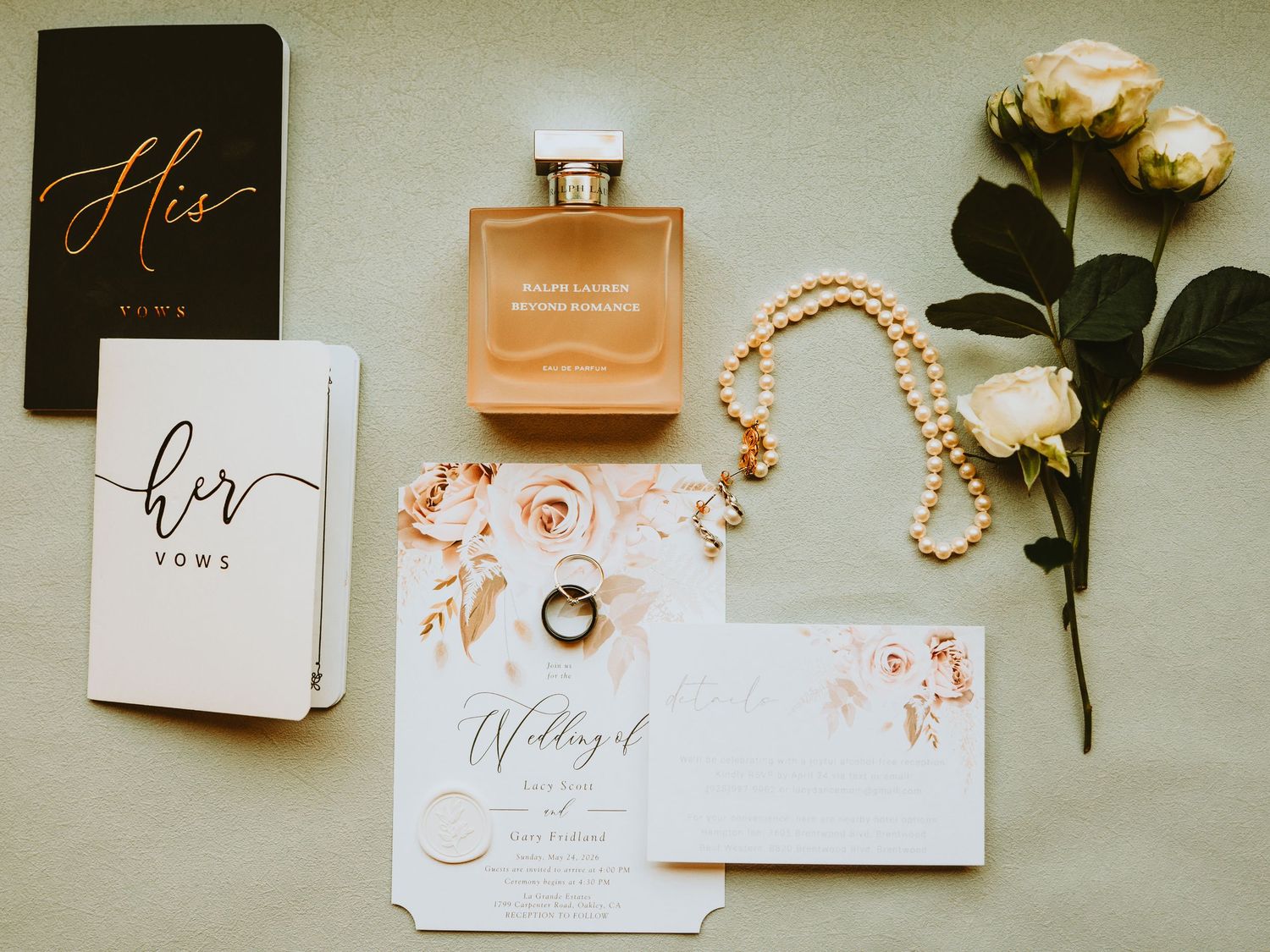 Wedding flat lay featuring vow books, perfume, pearl necklace, rings, floral invitation, and roses on neutral background.