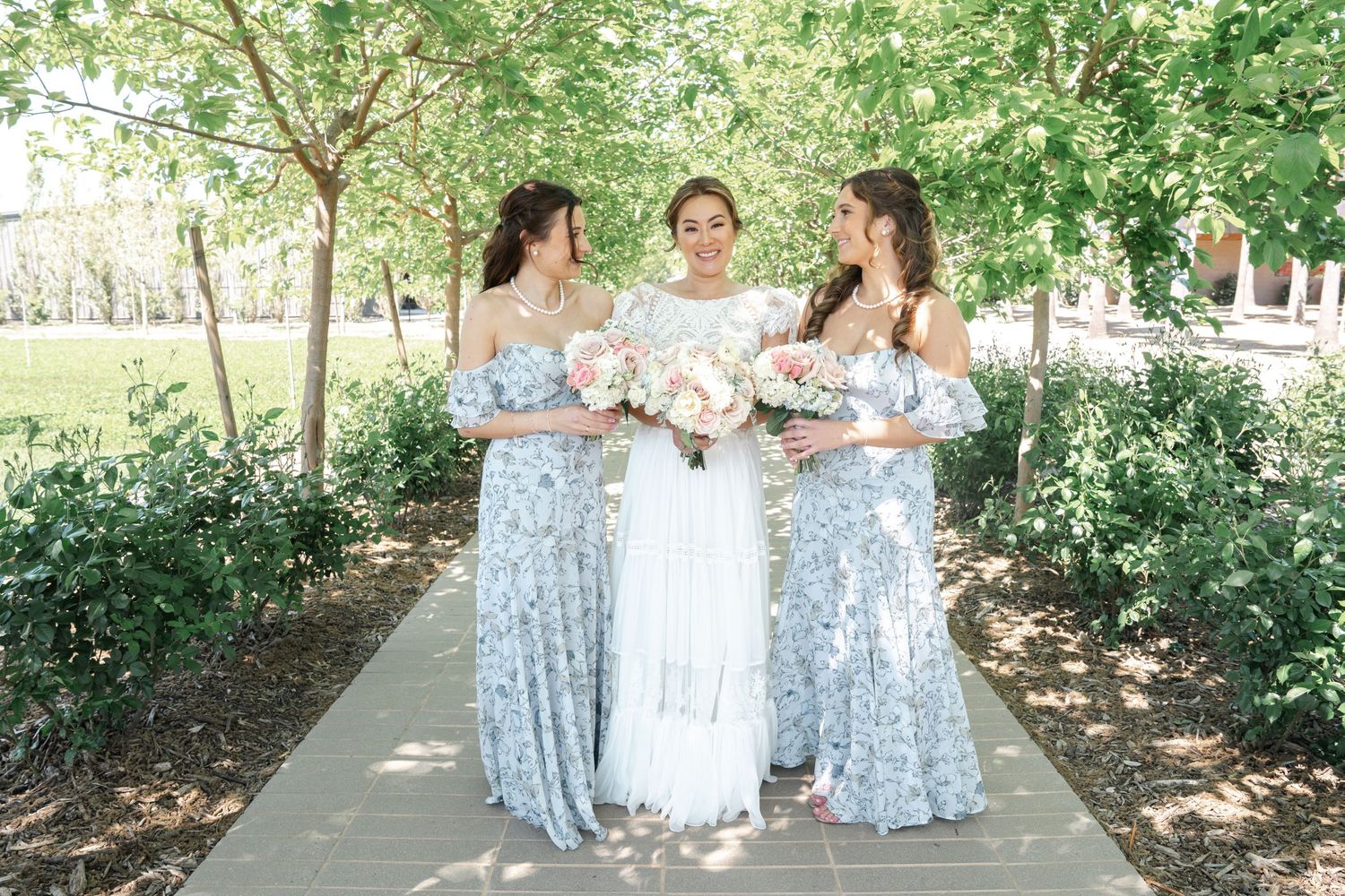 Three women in elegant dresses pose together on a garden path holding floral bouquets.