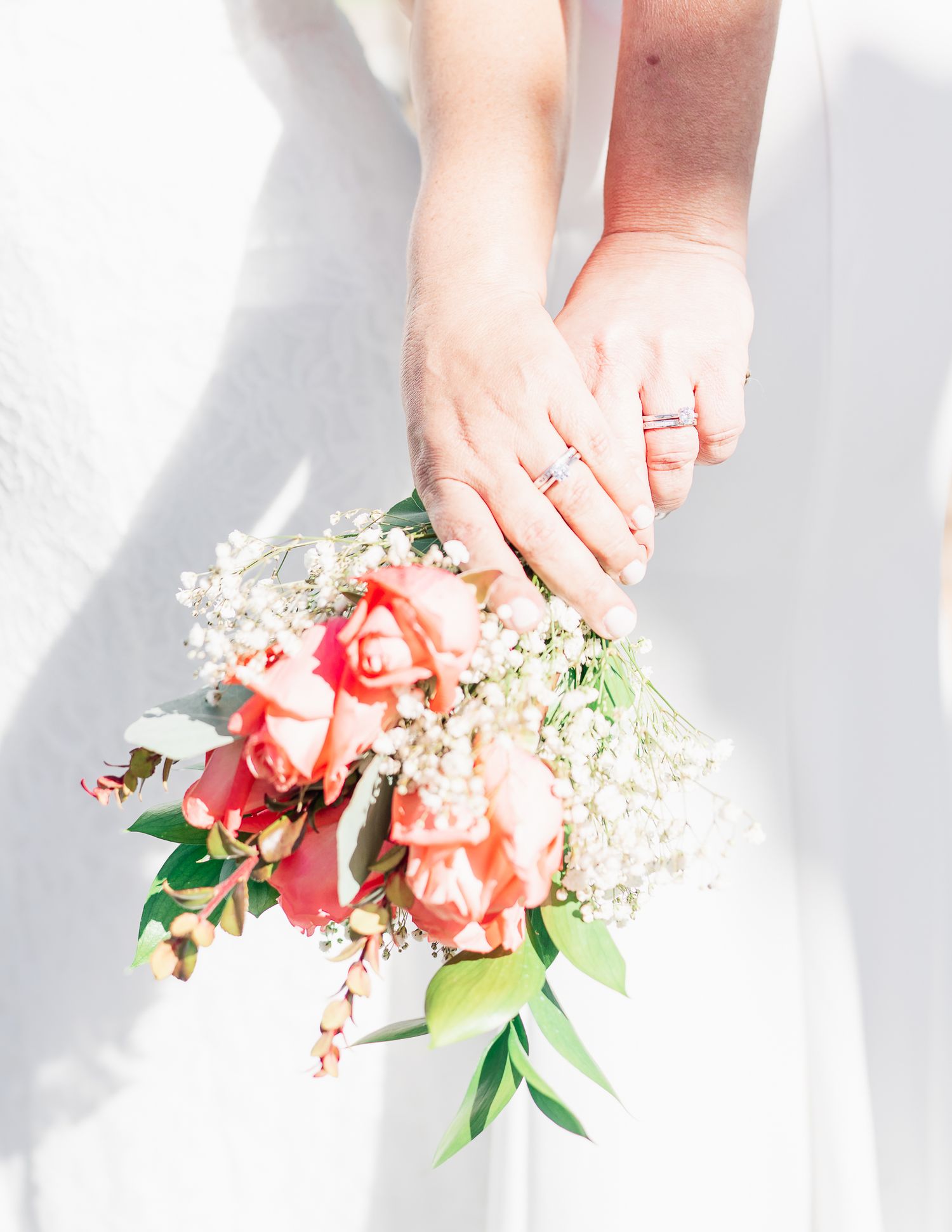 Close-up of two hands wearing wedding rings holding a bouquet of peach roses and baby's breath.