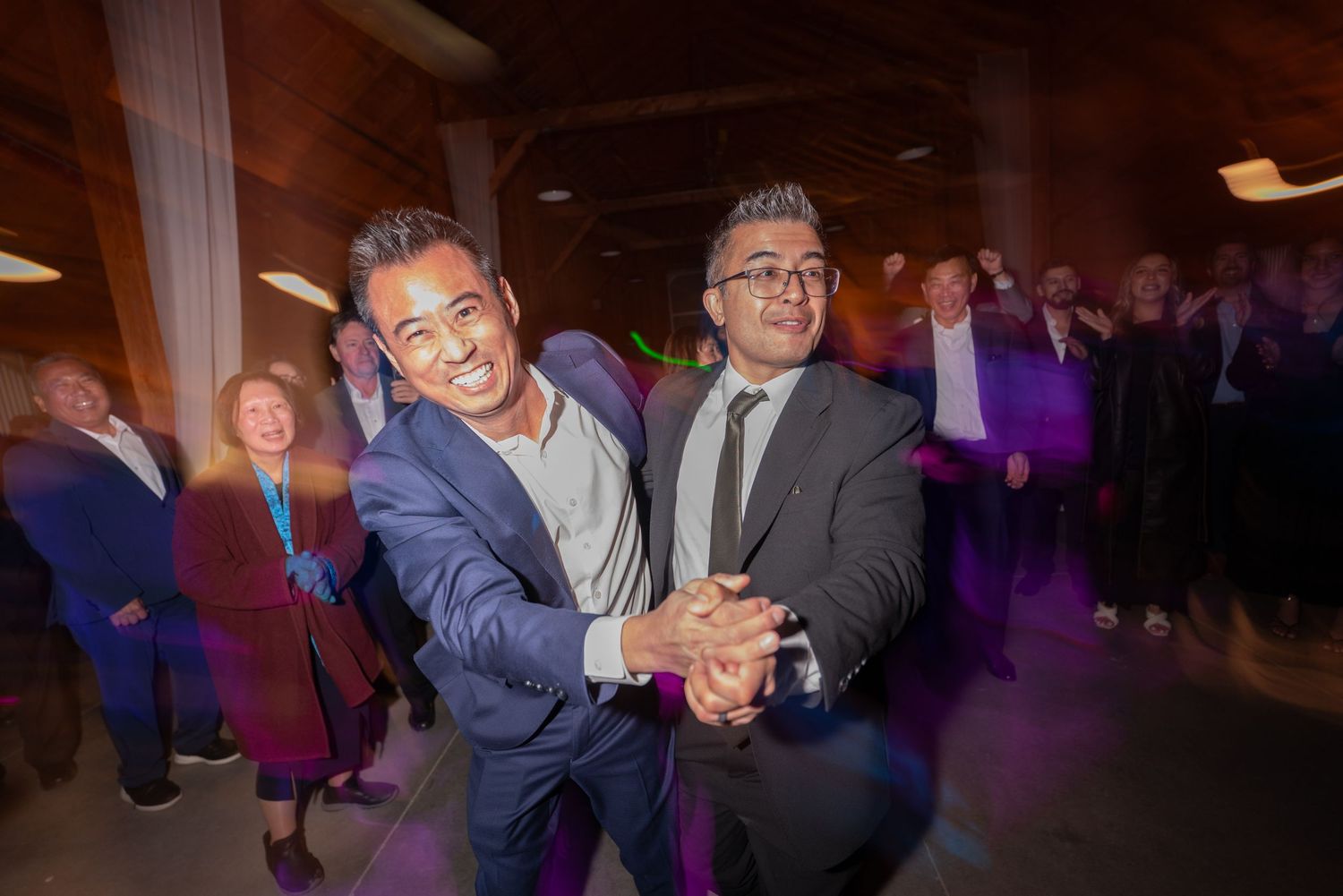 Two men in suits smiling and dancing together at a lively evening event with guests in the background.