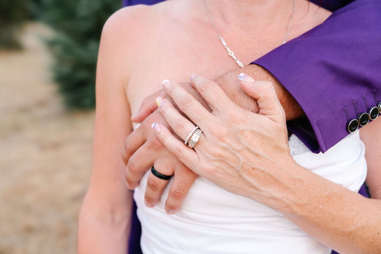 Close-up of bride and groom hands embracing, showing wedding ring, white dress, and purple suit jacket.