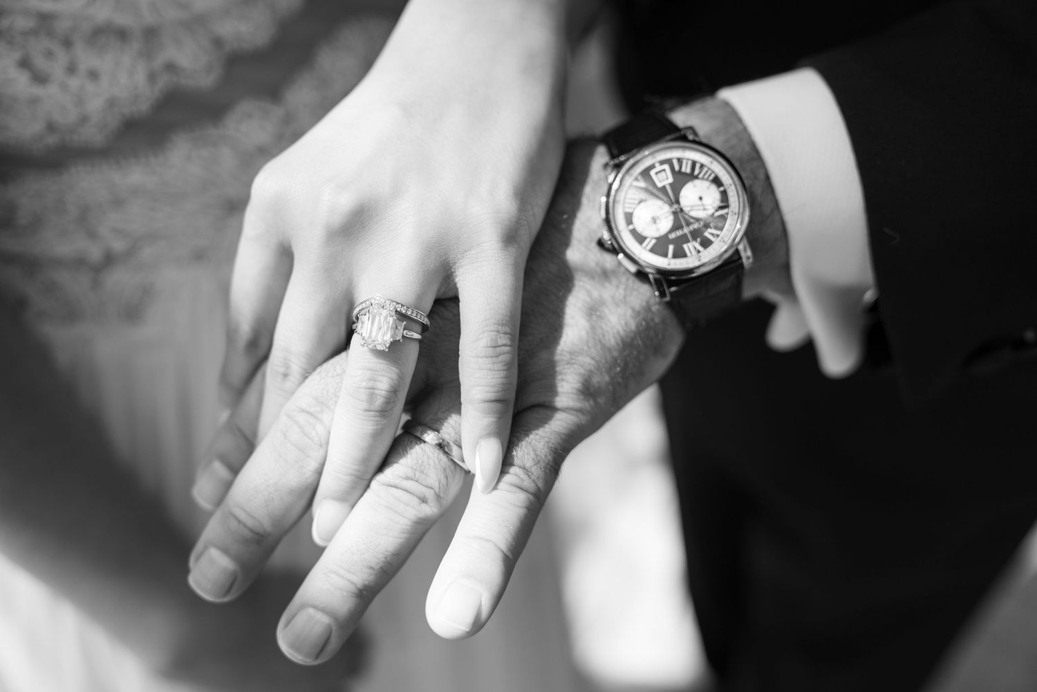 Black and white photo of bride and groom hands showing wedding rings, groom wearing luxury chronograph watch.
