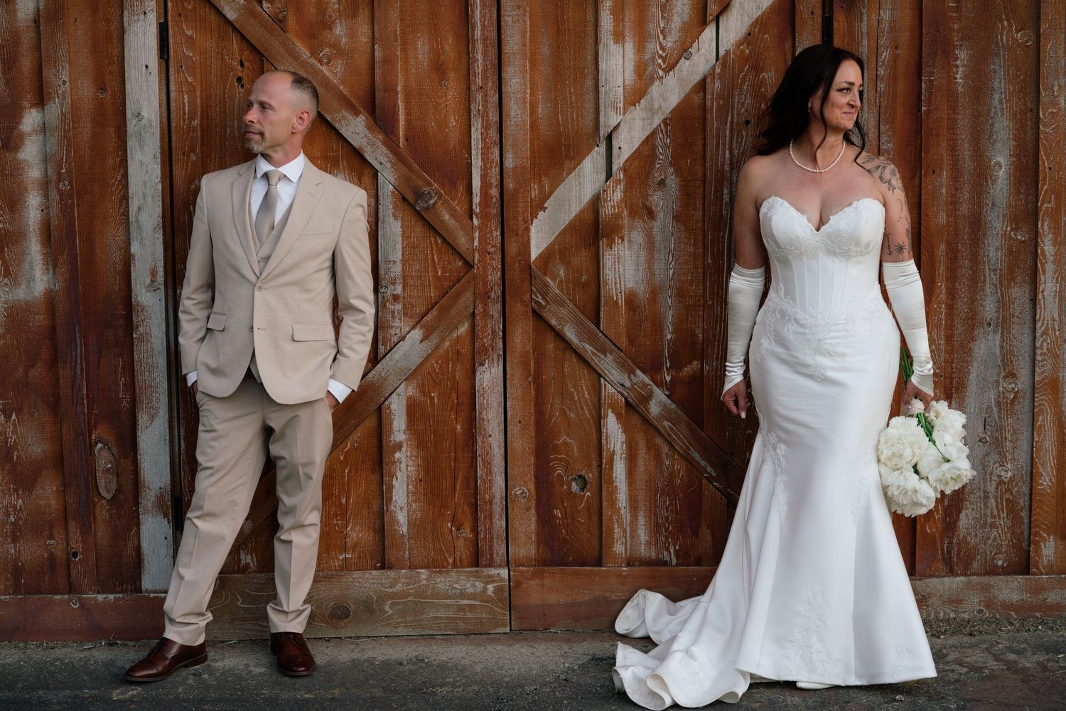 Bride in white gown with gloves and groom in tan suit pose separately before rustic wooden barn doors.