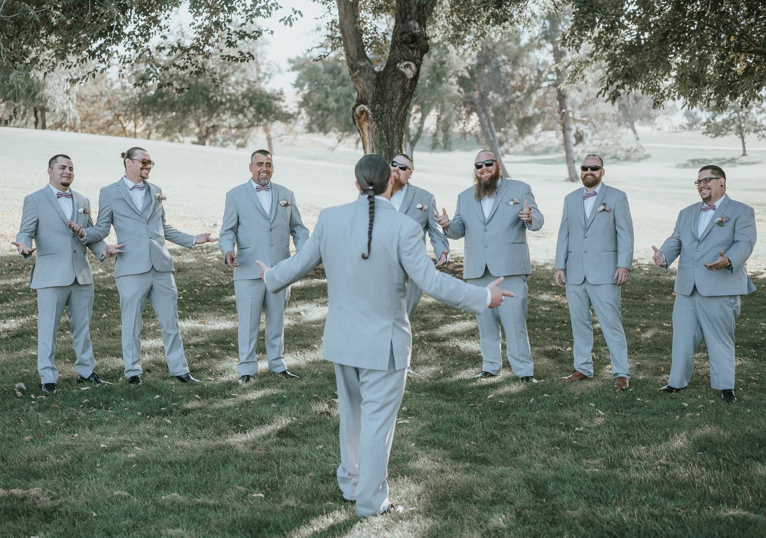 A groom in a gray suit faces his groomsmen in matching attire on a green lawn during a wedding.