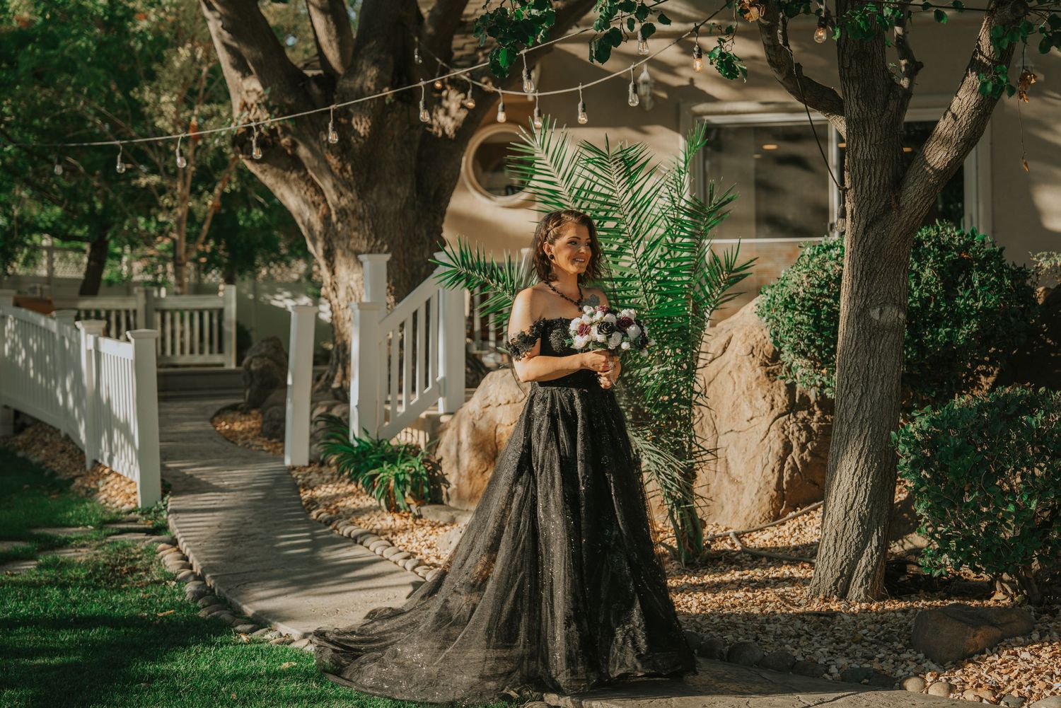 Woman in black off-shoulder ball gown holding bouquet stands among trees in a garden setting.