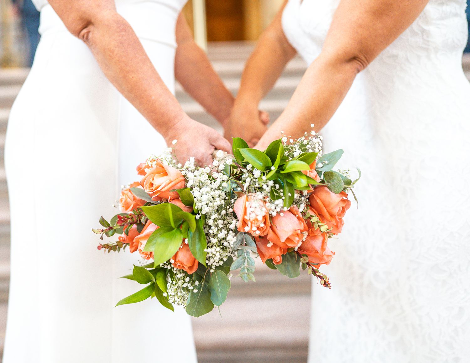 Two brides in white dresses holding matching coral rose and baby's breath bouquets at their wedding ceremony.