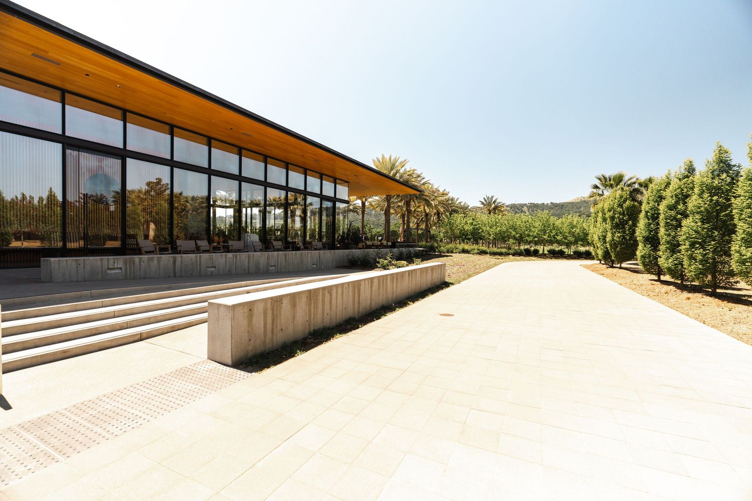 Modern pavilion building with floor-to-ceiling windows, wooden canopy roof, and stone steps surrounded by lush greenery.