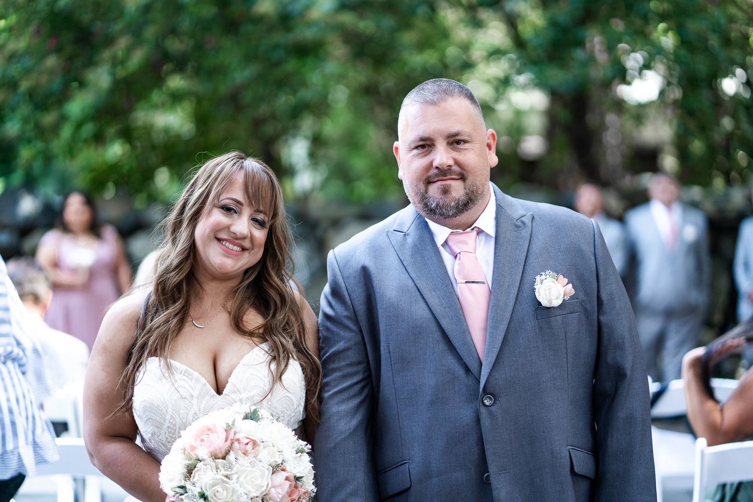 Smiling bride and groom pose together outdoors during wedding ceremony, bride holding pink floral bouquet.