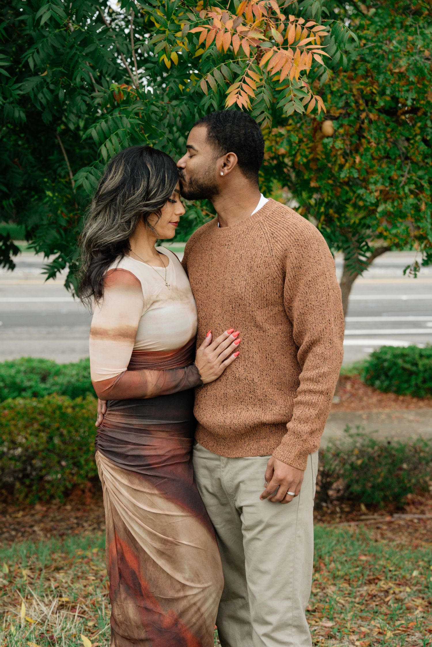 A couple shares an intimate moment among autumn foliage wearing a brown sweater and beige dress.