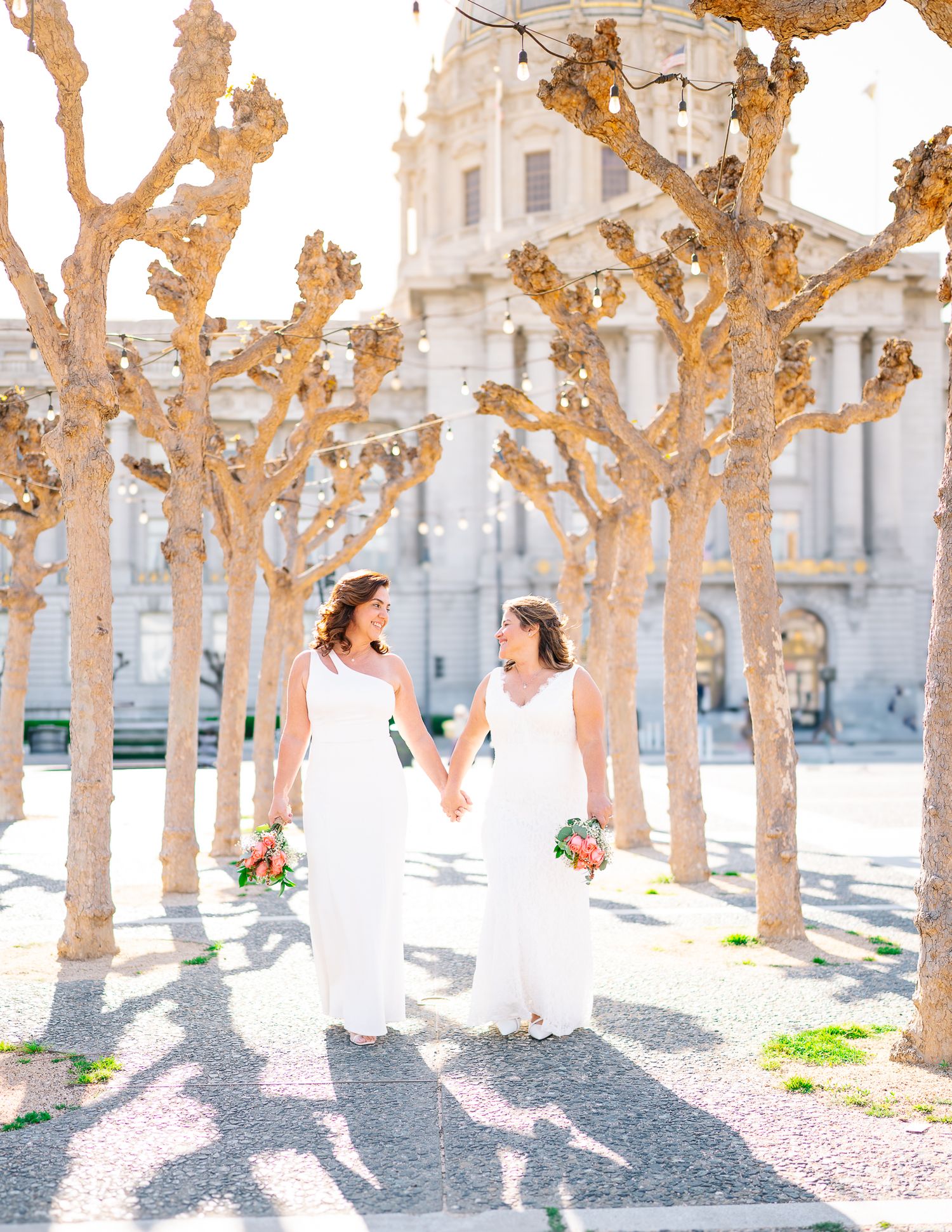 Two brides in white gowns hold hands and laugh among golden trees outside a grand building on their wedding day.