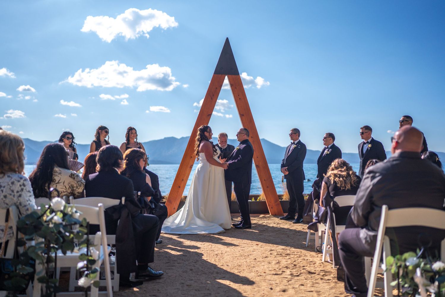 Outdoor wedding ceremony under a wooden A-frame arch with mountain lake views on a sunny day.
