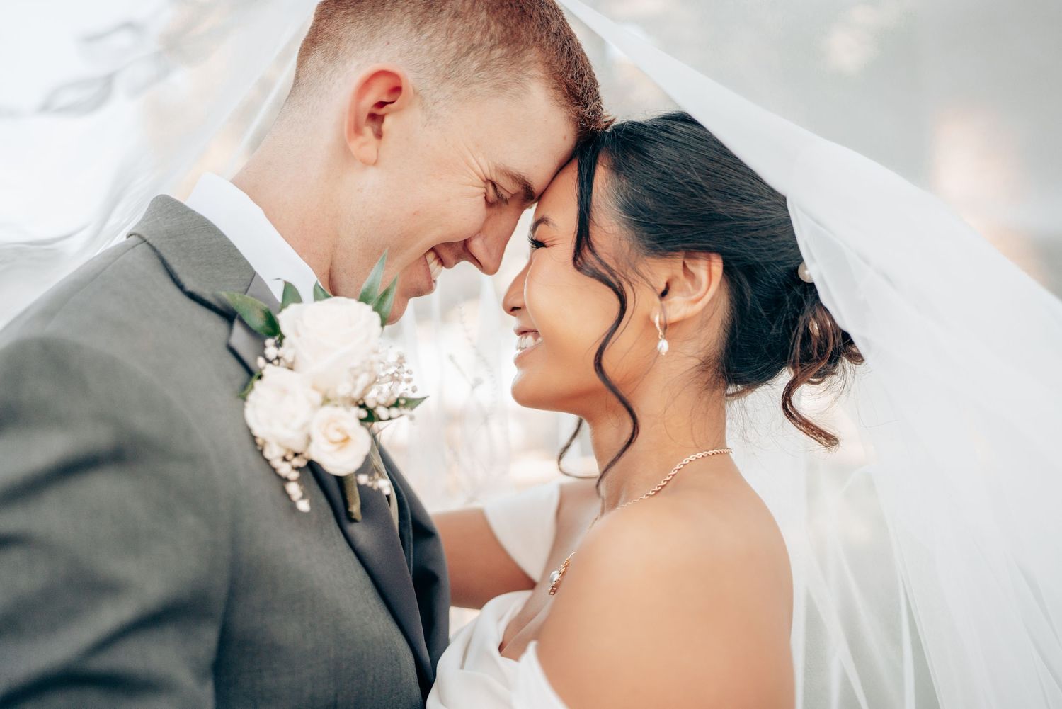A bride and groom touch foreheads tenderly under a white veil on their wedding day.