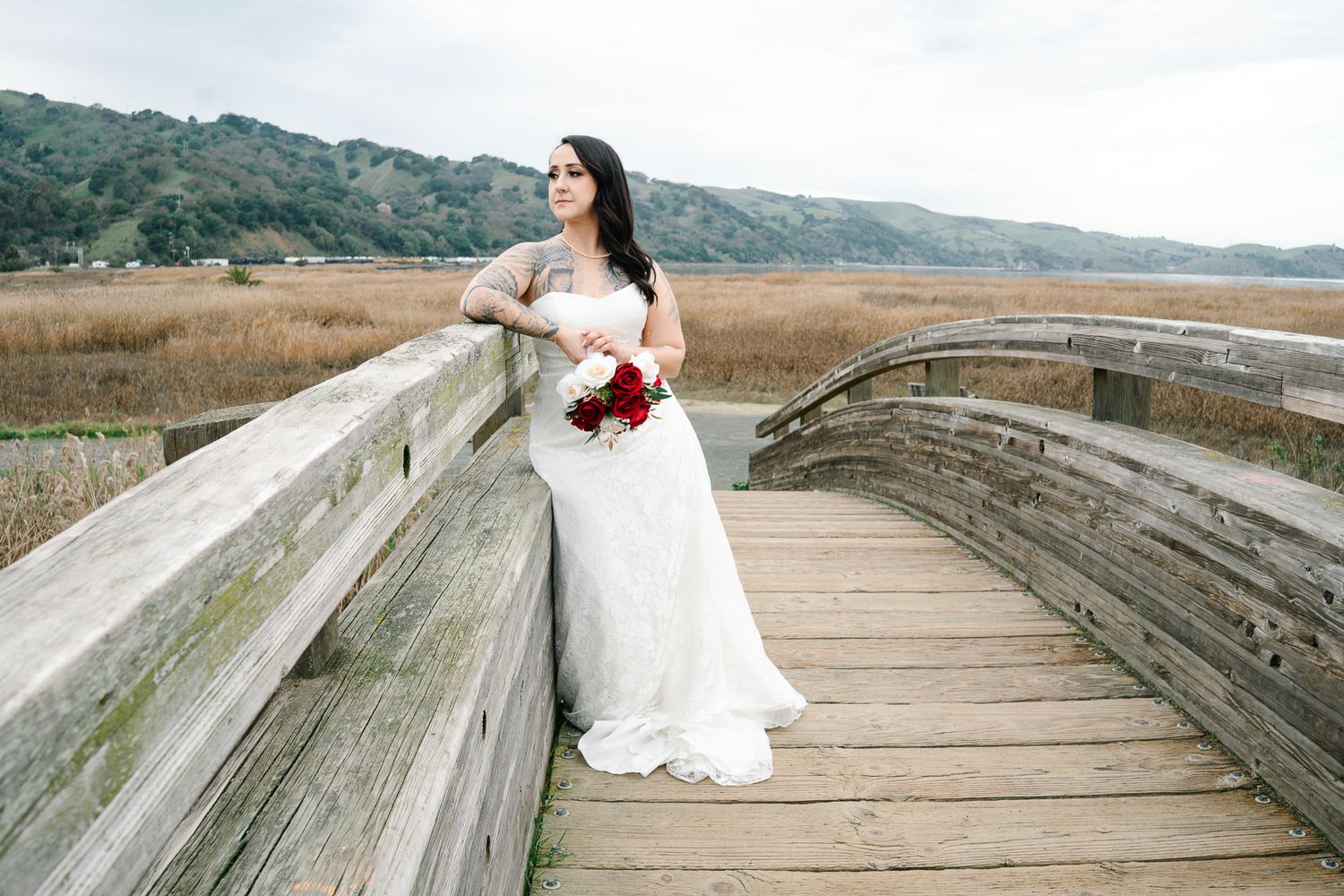 Bride in white lace gown holding red bouquet on wooden boardwalk overlooking scenic marshland and hills.