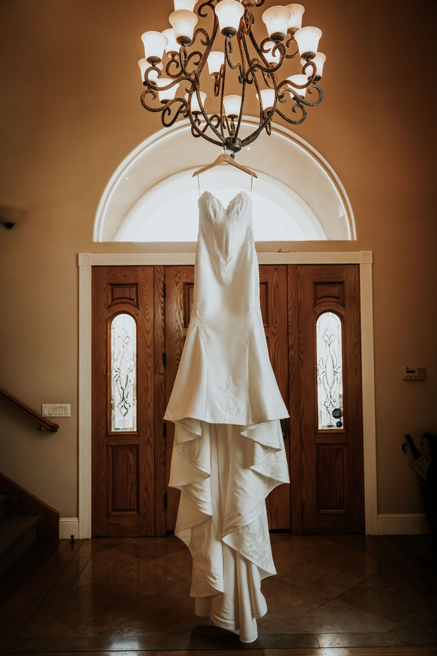 Elegant white mermaid wedding dress hanging from a chandelier in a grand foyer with wooden double doors.