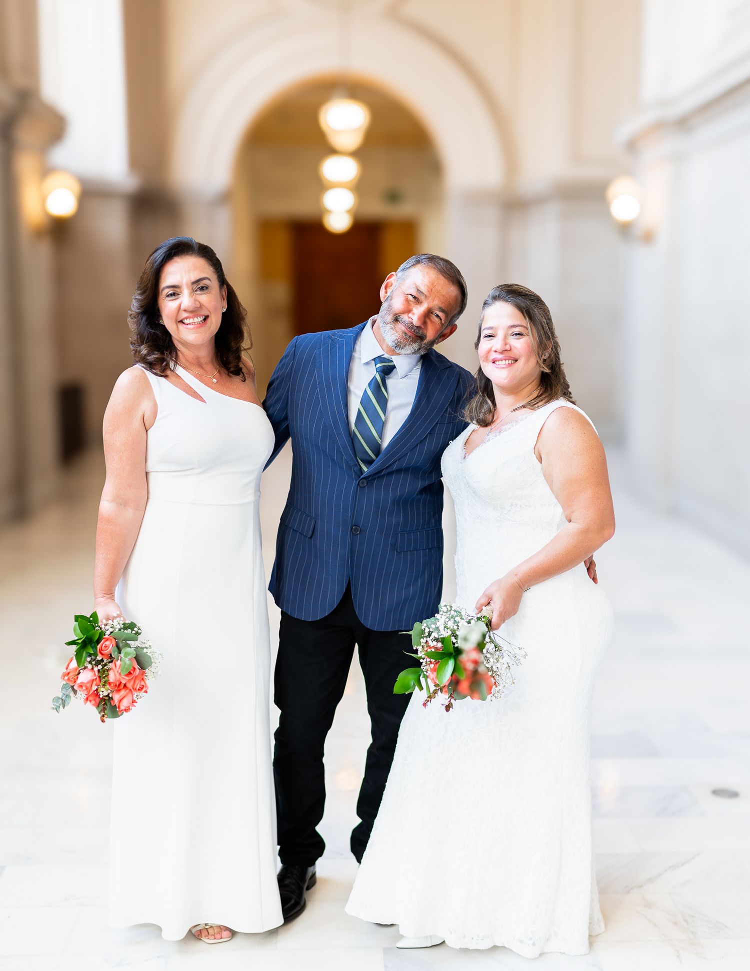 Two brides in white gowns holding bouquets pose with a man in a blue suit at an elegant wedding venue.