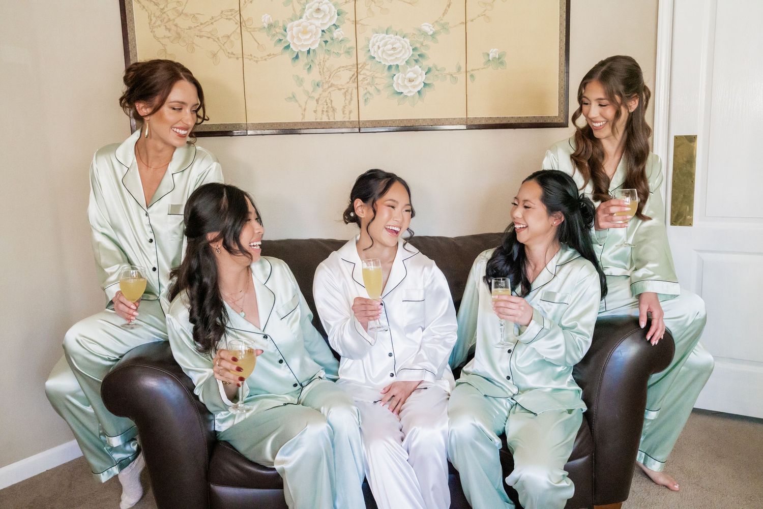 Bridesmaids in sage green satin pajamas laughing on a sofa holding champagne glasses during bridal getting ready party.