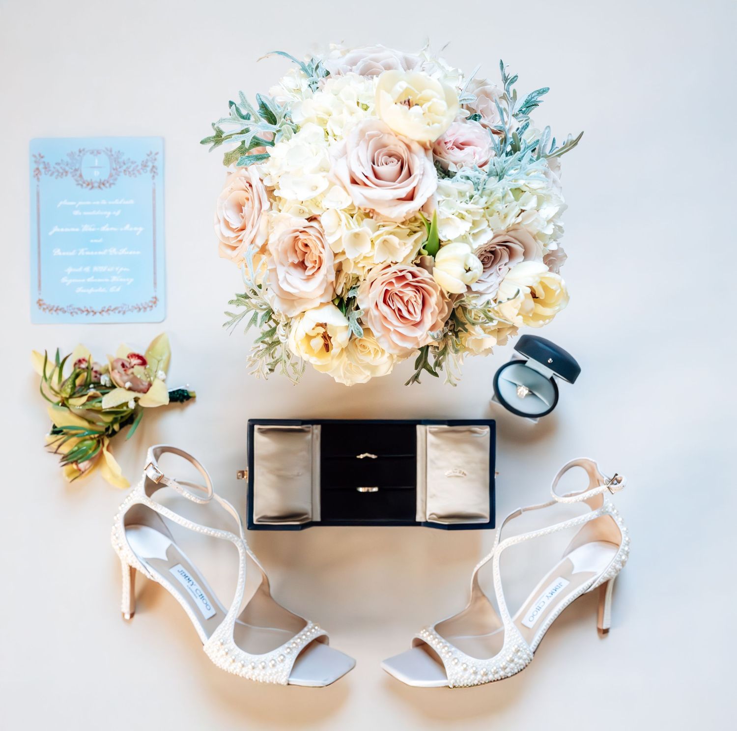 Elegant wedding flat lay with blush roses bouquet, white heels, ring box, boutonniere, and blue invitation on white background.