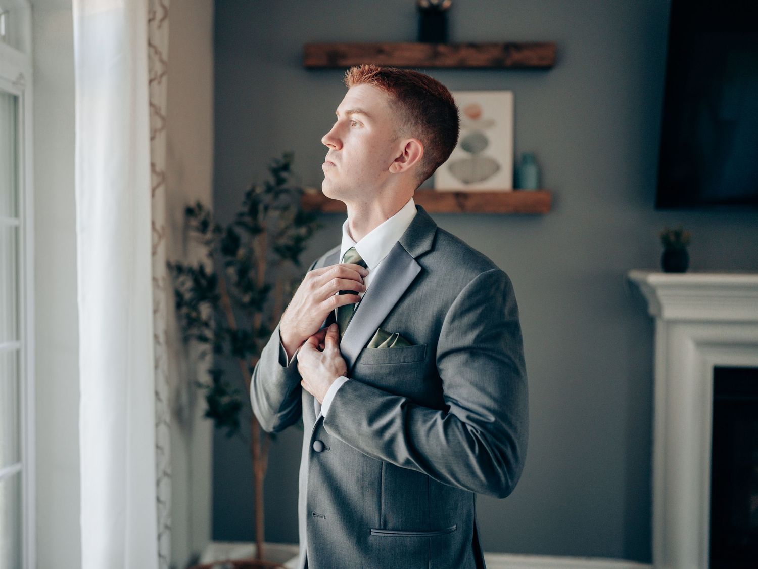 A well-dressed groom in a gray suit adjusts his tie while standing near a window in an elegant room.