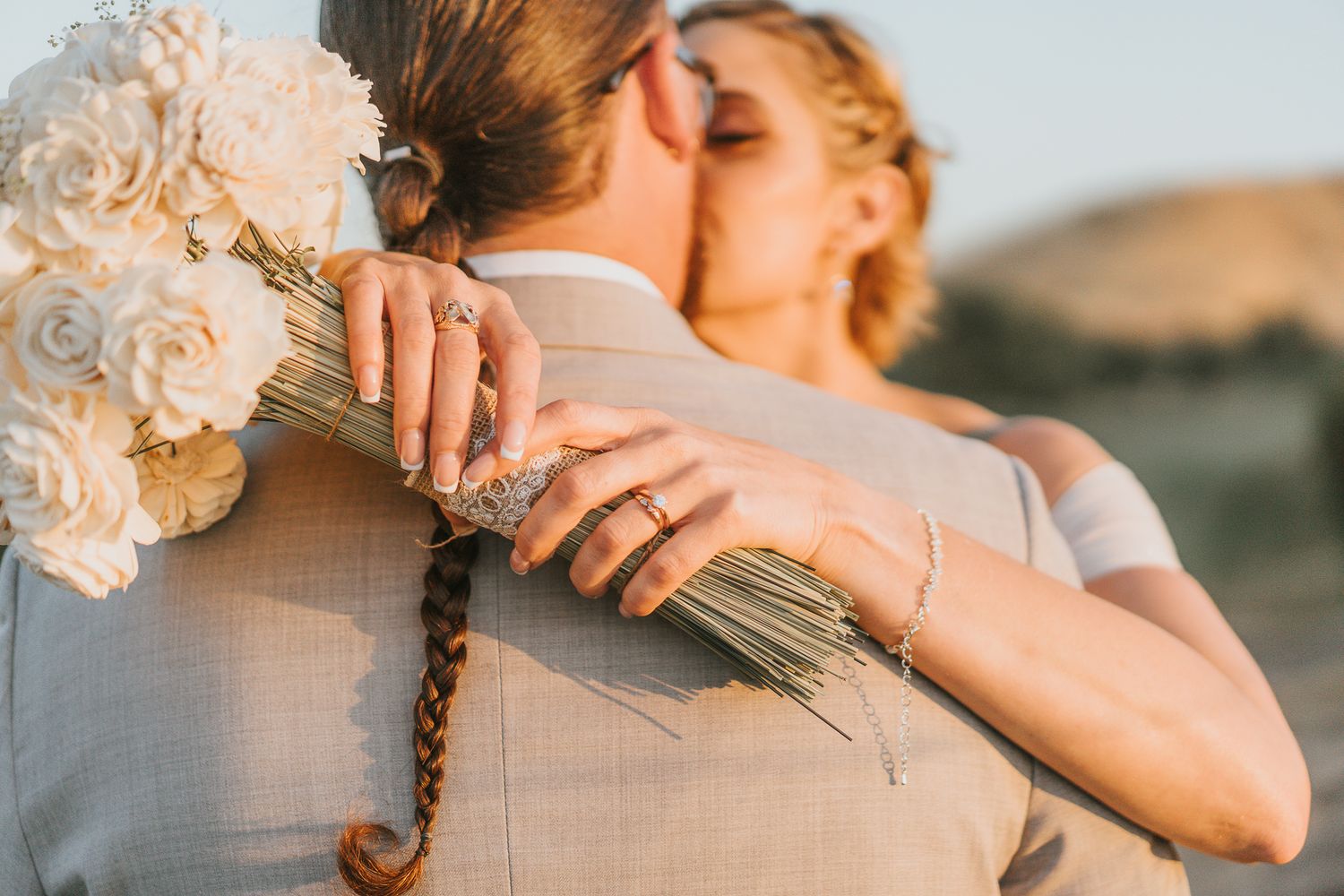 Bride and groom sharing a kiss, bride holding white rose bouquet, wearing gold rings, at golden hour outdoor wedding.