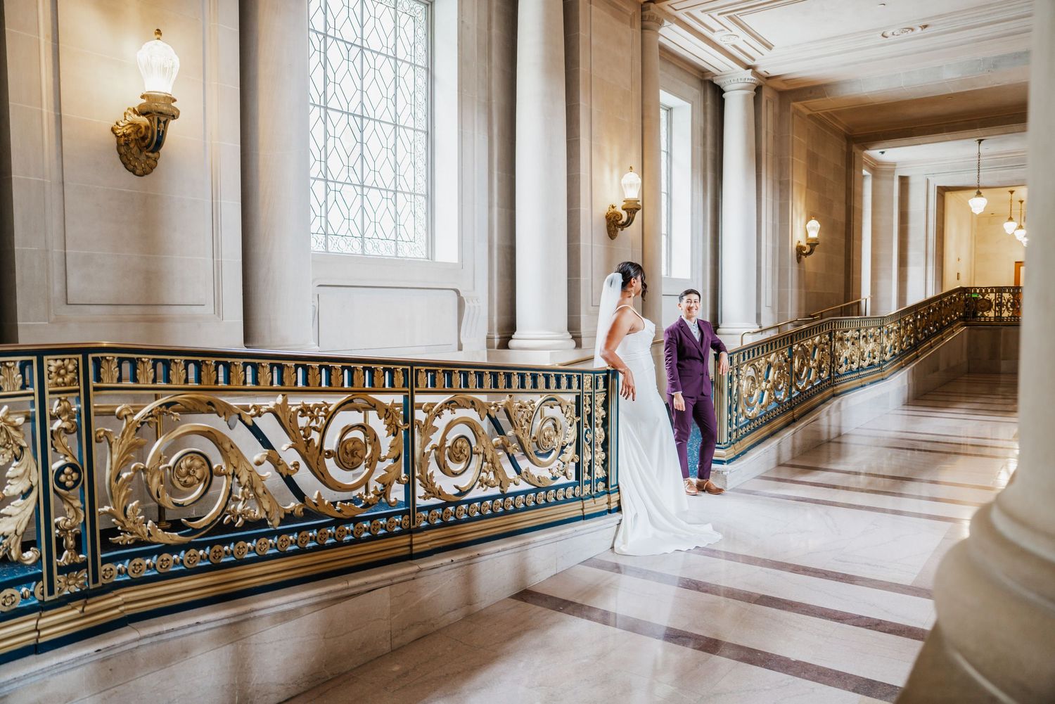 Bride and groom pose on ornate golden staircase railing inside San Francisco City Hall during wedding ceremony.