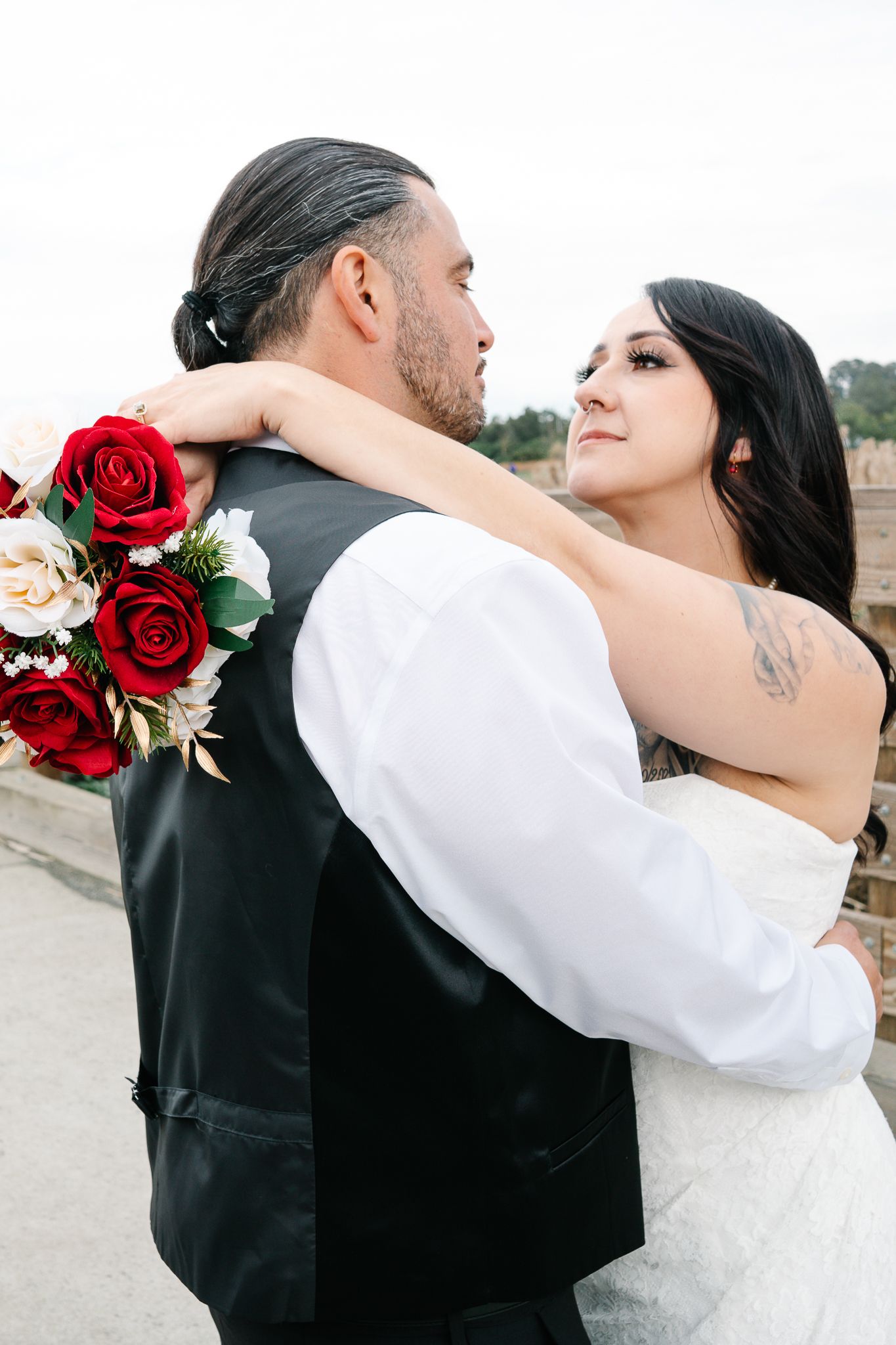 Bride and groom embracing, with red and white rose bouquet, at outdoor wedding ceremony.