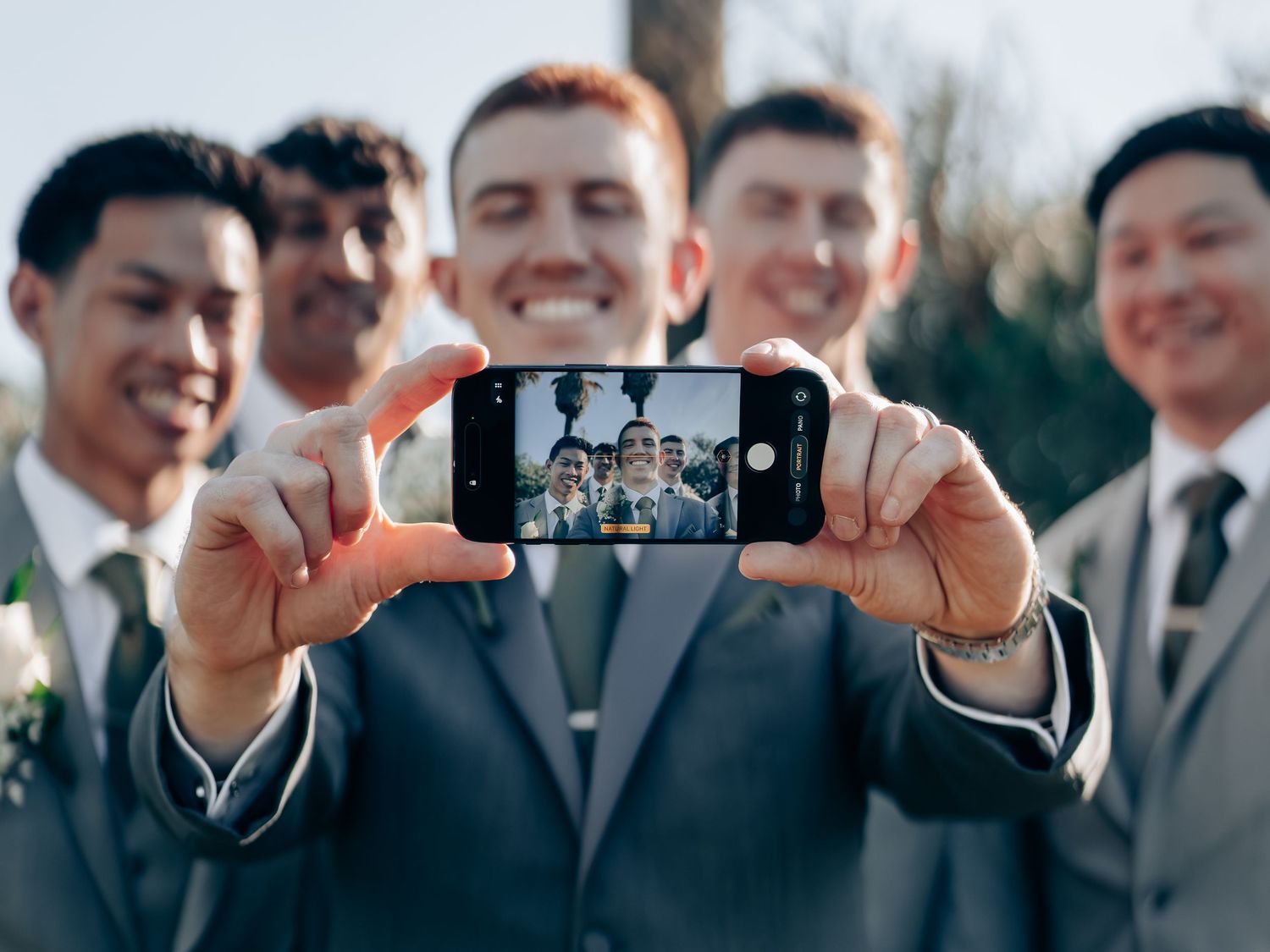 Groomsmen in gray suits take a fun selfie, with the groom holding a smartphone showing their reflection at a wedding.