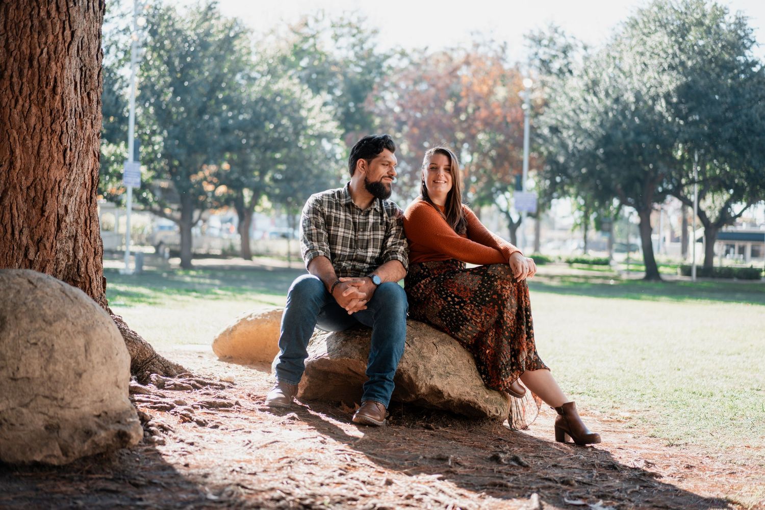 Couple sitting together near large tree trunk in sunny park setting.